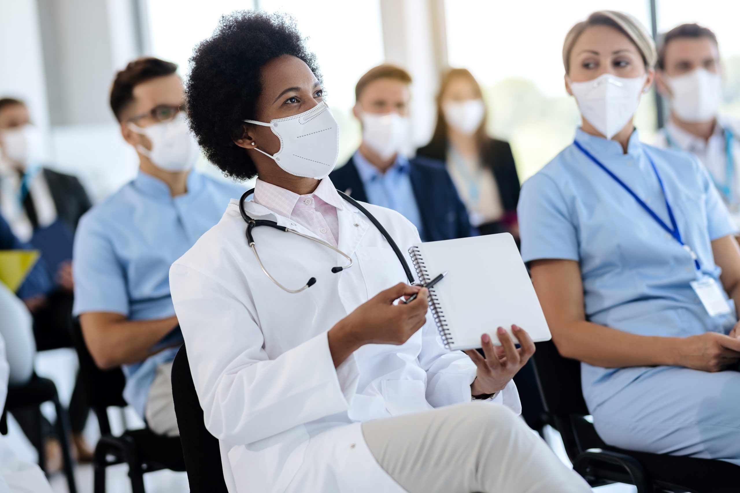 Happy black doctor pointing at her notebook while being on a seminar with group of colleagues during COVID-19 pandemic.